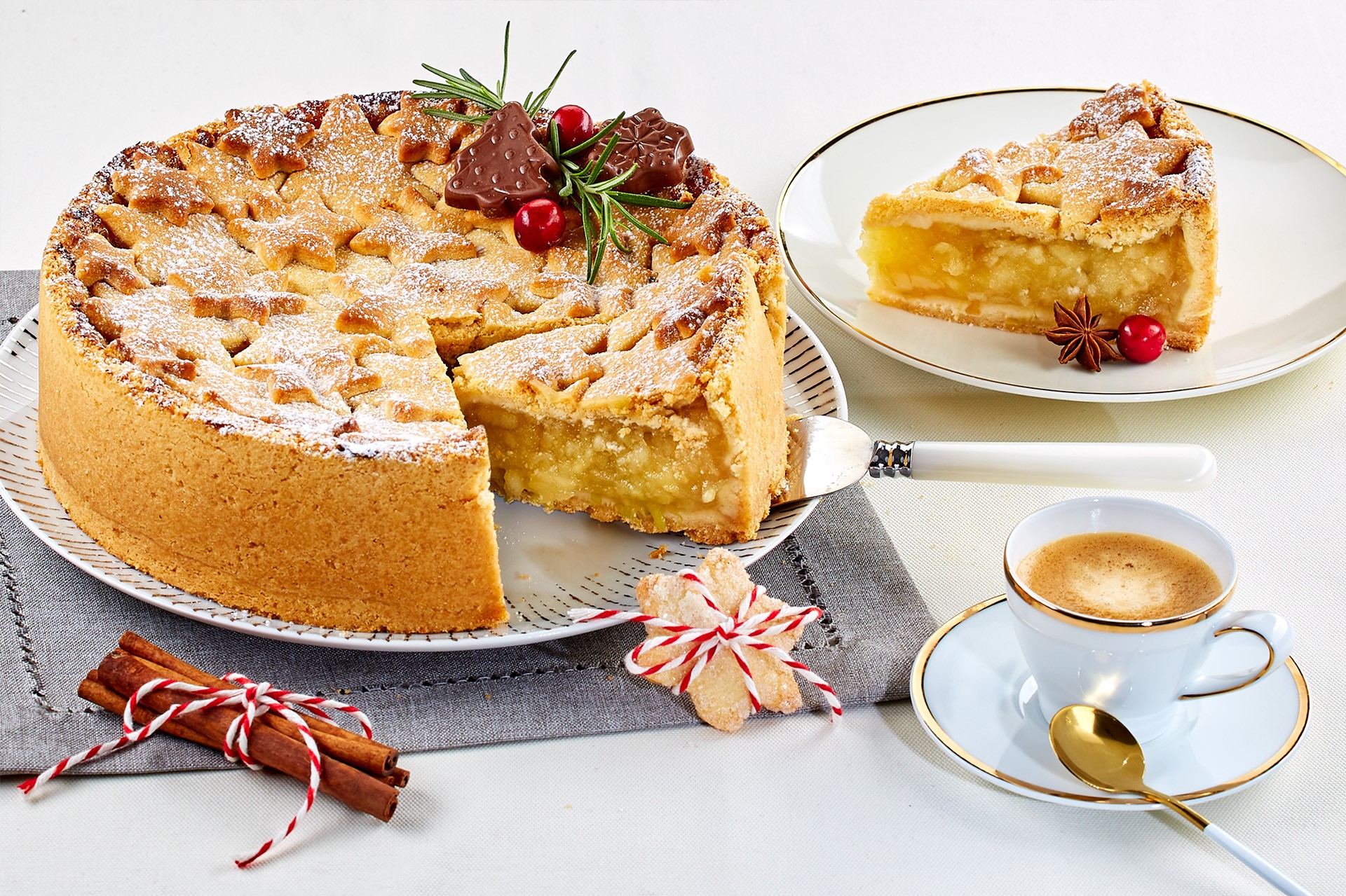 A golden-brown lattice apple pie topped with powdered sugar is partially sliced on a patterned plate, accompanied by a plate with a slice of the pie, a sprig of rosemary, star anise, and chocolate decorations, next to a cinnamon stick and a cup of espresso on a white table.