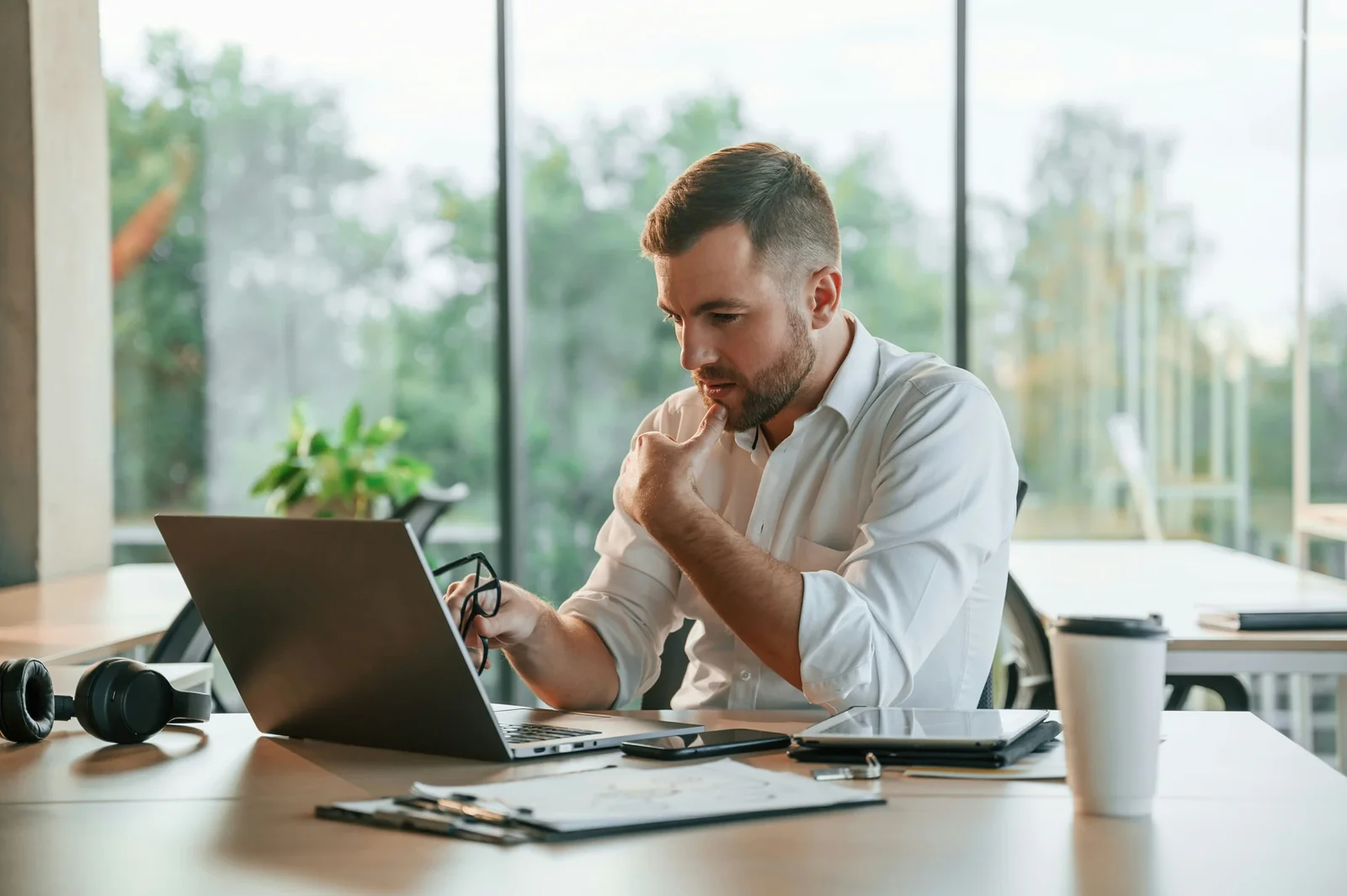 Professional man in white shirt working at laptop in modern office with large windows overlooking green landscape.