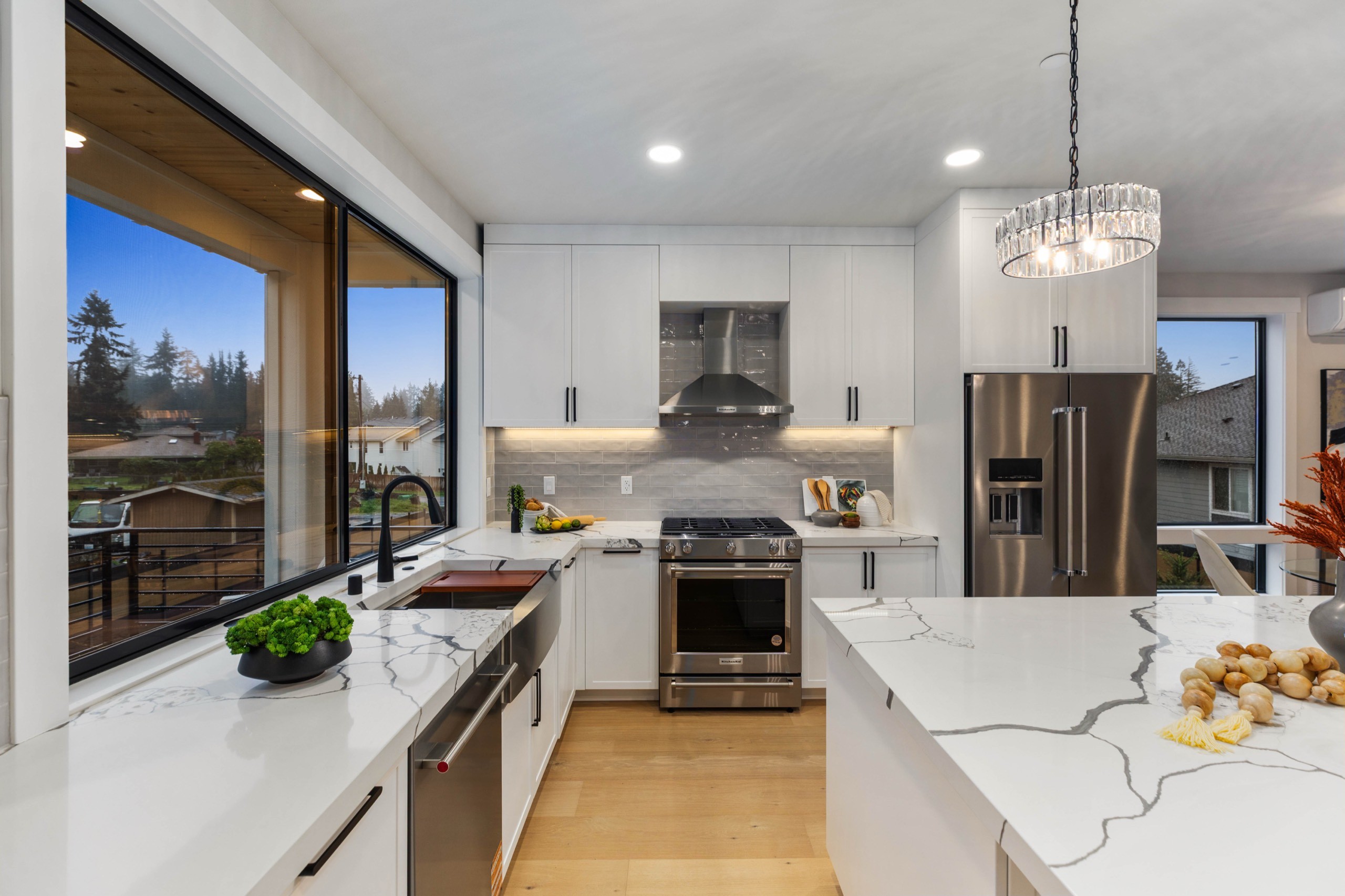 Spacious luxury kitchen with white shaker cabinets, dramatic waterfall quartz island, stainless steel appliances, designer chandelier, and large windows with neighbourhood views