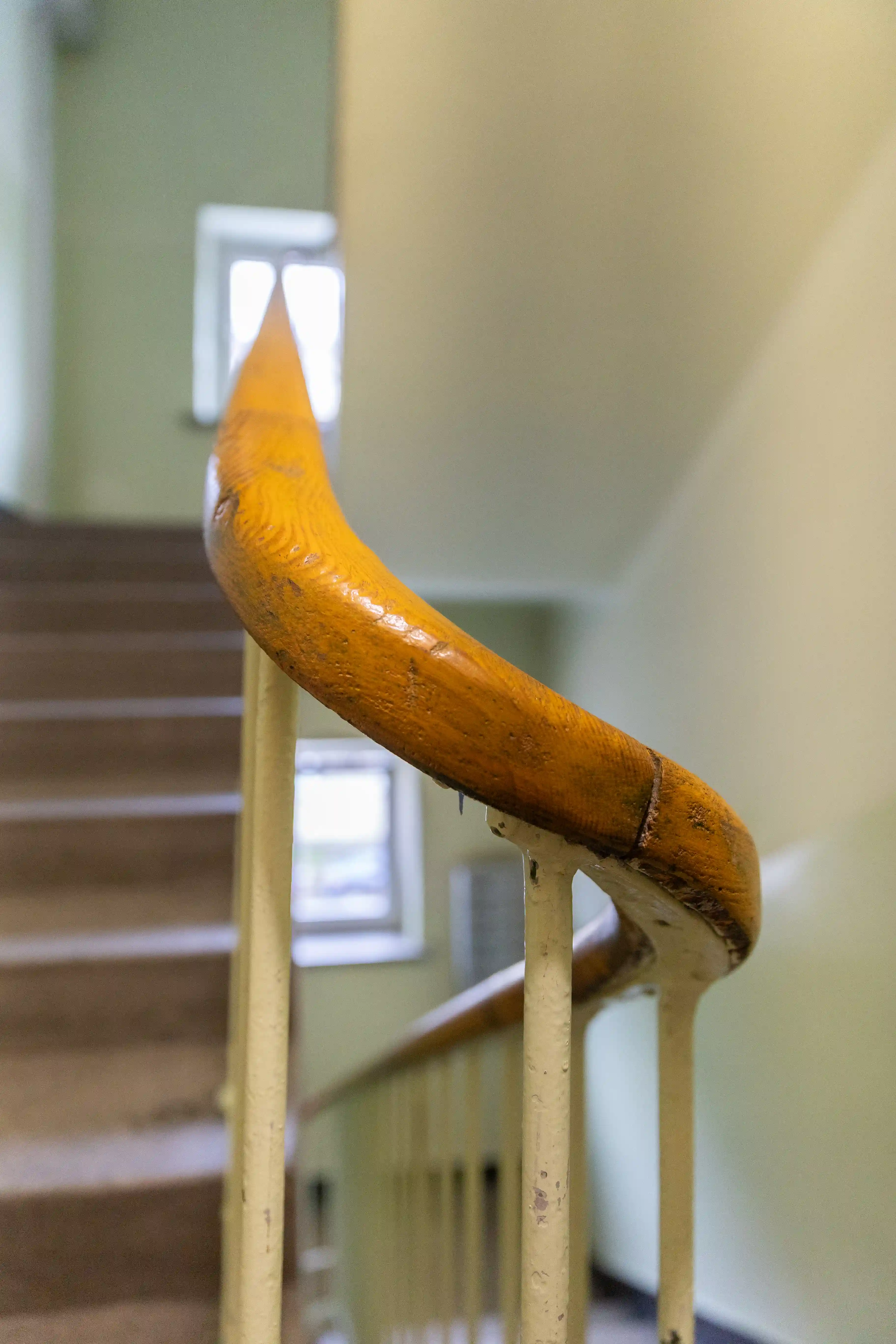 Drewniany uchwyt poręczy klatki schodowej w budynku mieszkalnym. W tle schody oraz małe okna. 