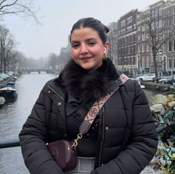 Smiling young woman with curly hair sitting at a laptop, in front of a teal wall.
