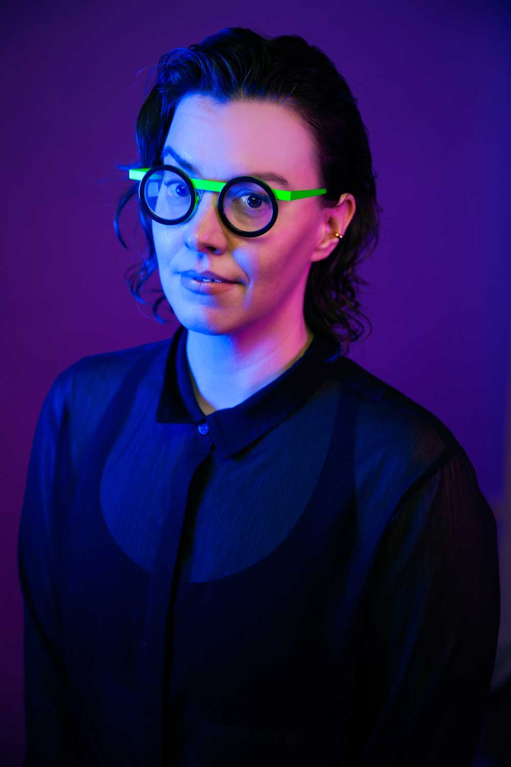 Margot stands on a transparent plastic mural with colorful shapes. They have short wavy hair and is white