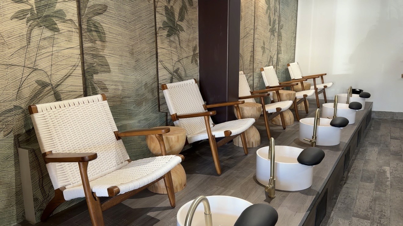 A serene and elegant spa space with wooden reclining chairs and white cushions. Golden sunlight filters through a cascading water feature, creating a peaceful atmosphere. Tropical plants and candles add to the relaxing ambiance.