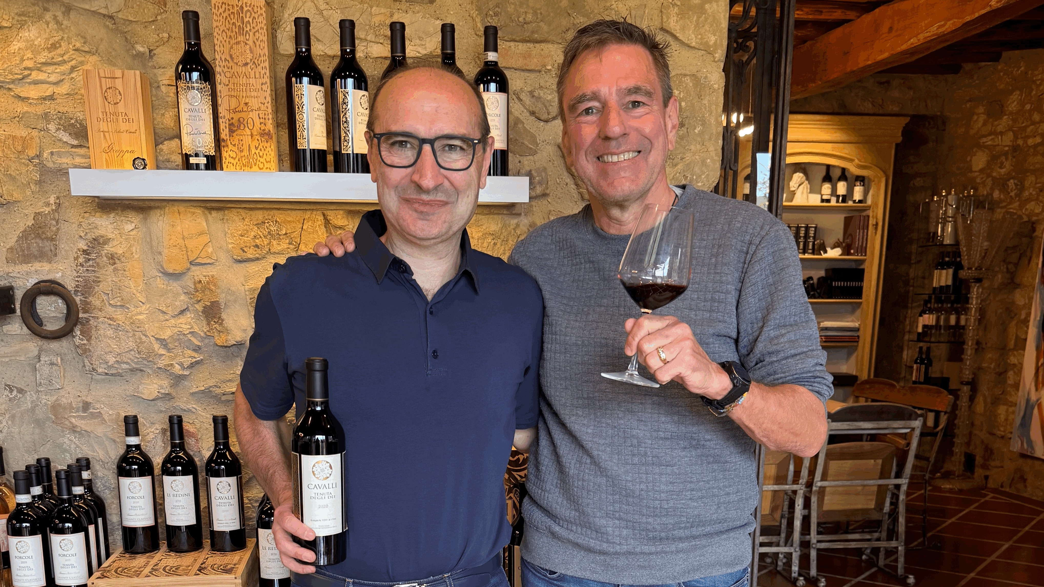 Two men stand together in the stone tasting room, one holding a bottle of 2020 Cavalli and the other holding a glass of red wine.