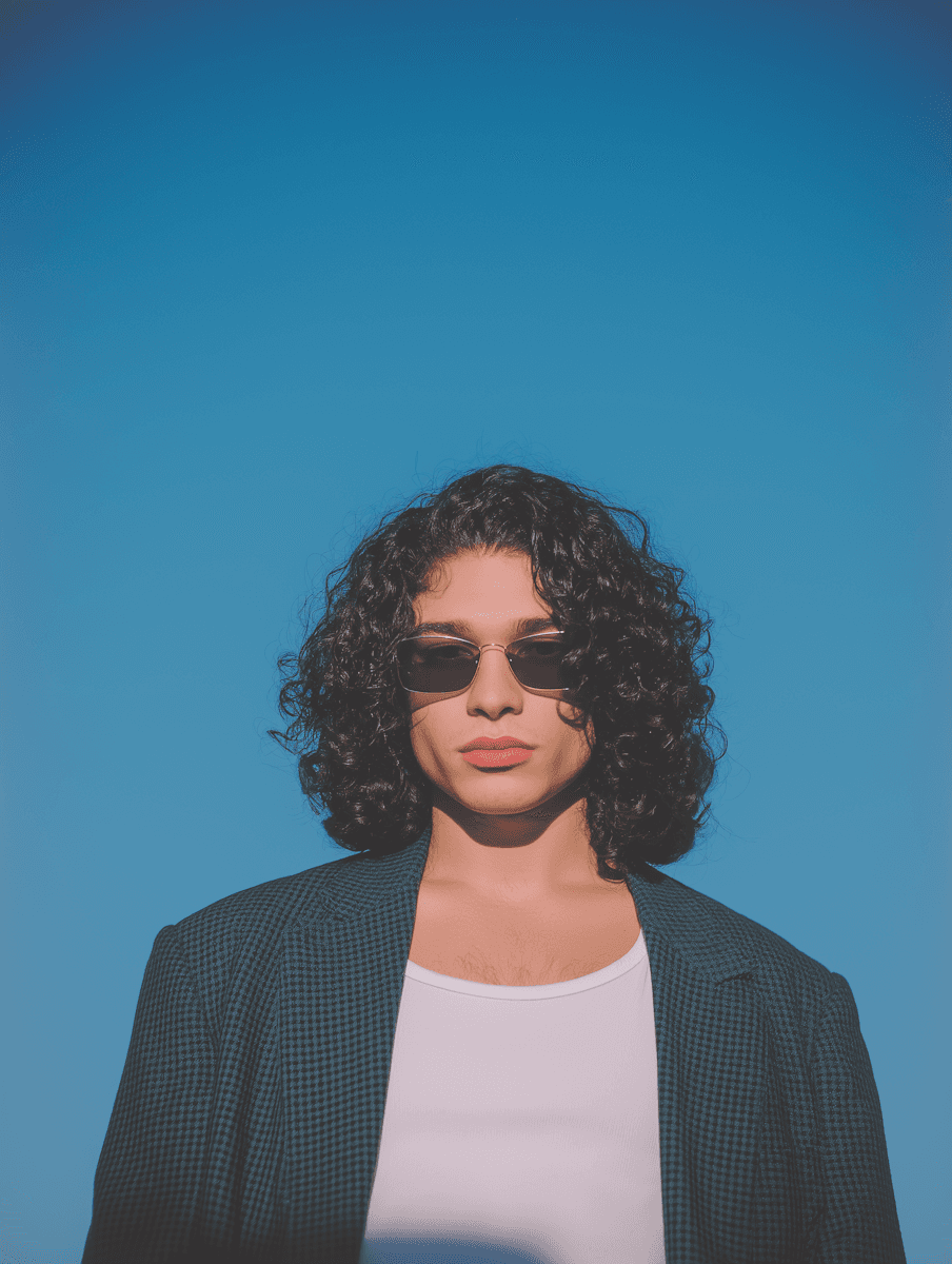 Stylish individual with curly hair and sunglasses against a bright blue sky, wearing a white tank top and a dark blazer.