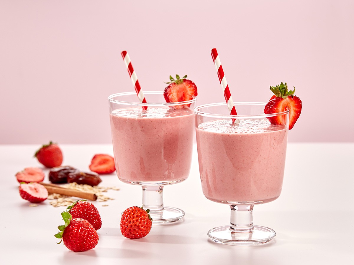 Two glasses of creamy strawberry smoothies, garnished with fresh strawberries and red striped straws, are surrounded by whole and sliced strawberries on a light pink background.