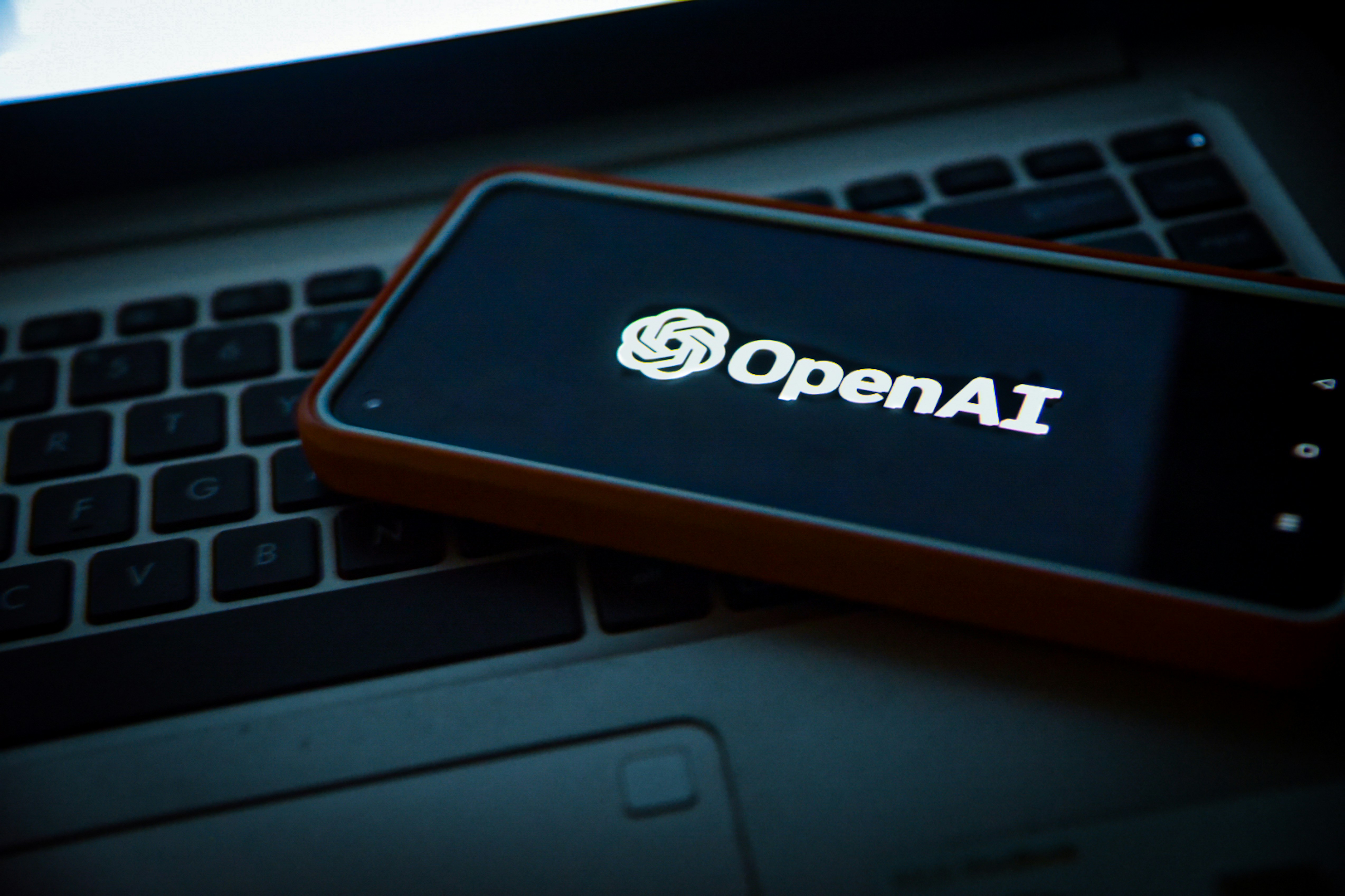 An open AI logo in white is visibly displaced on a phone screen. The phone is placed on the keyboard of a laptop. 