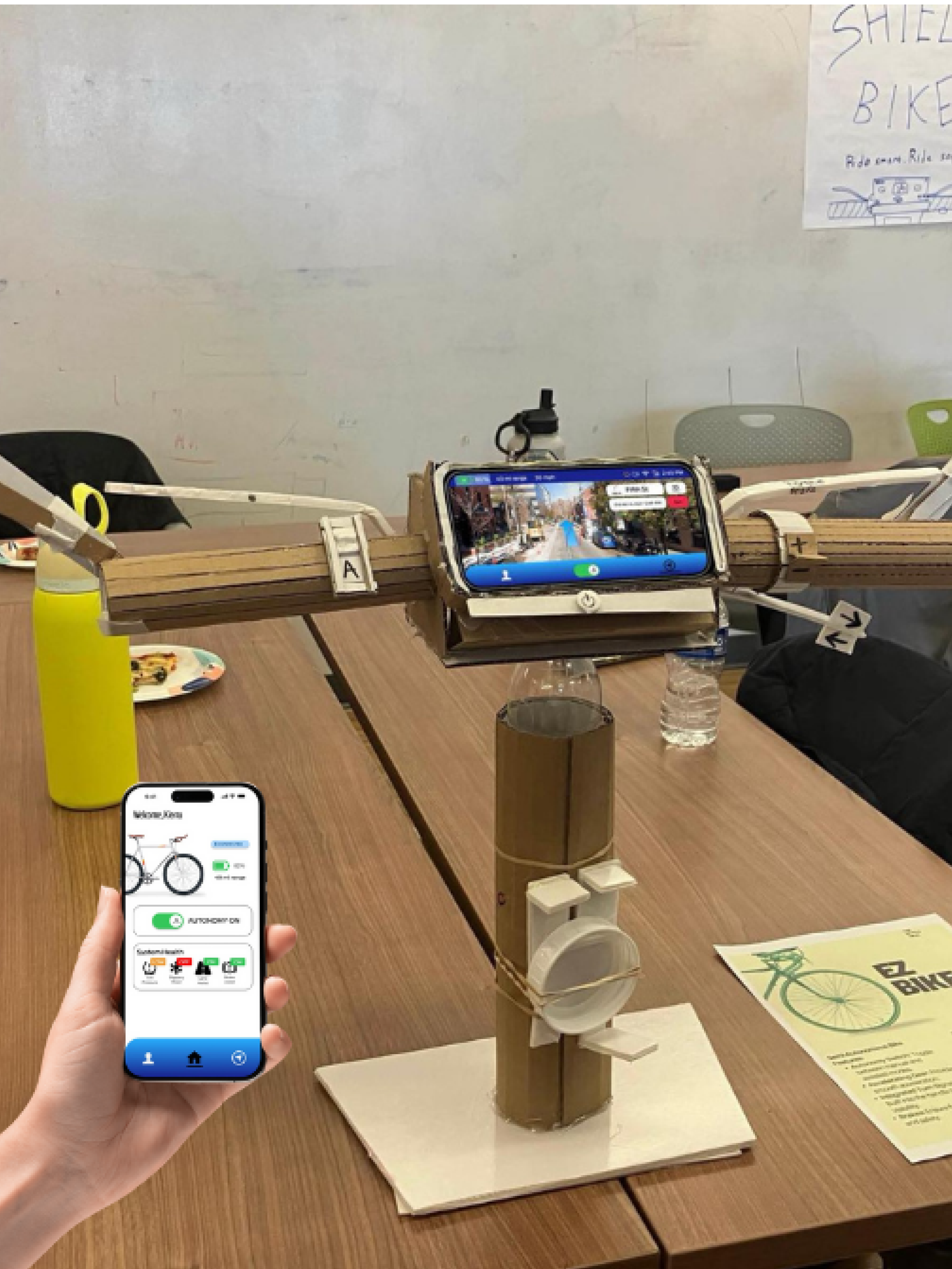 Overview of bike controls made of cardboard and a hand to the side holding a phone with the home screen of the mobile app