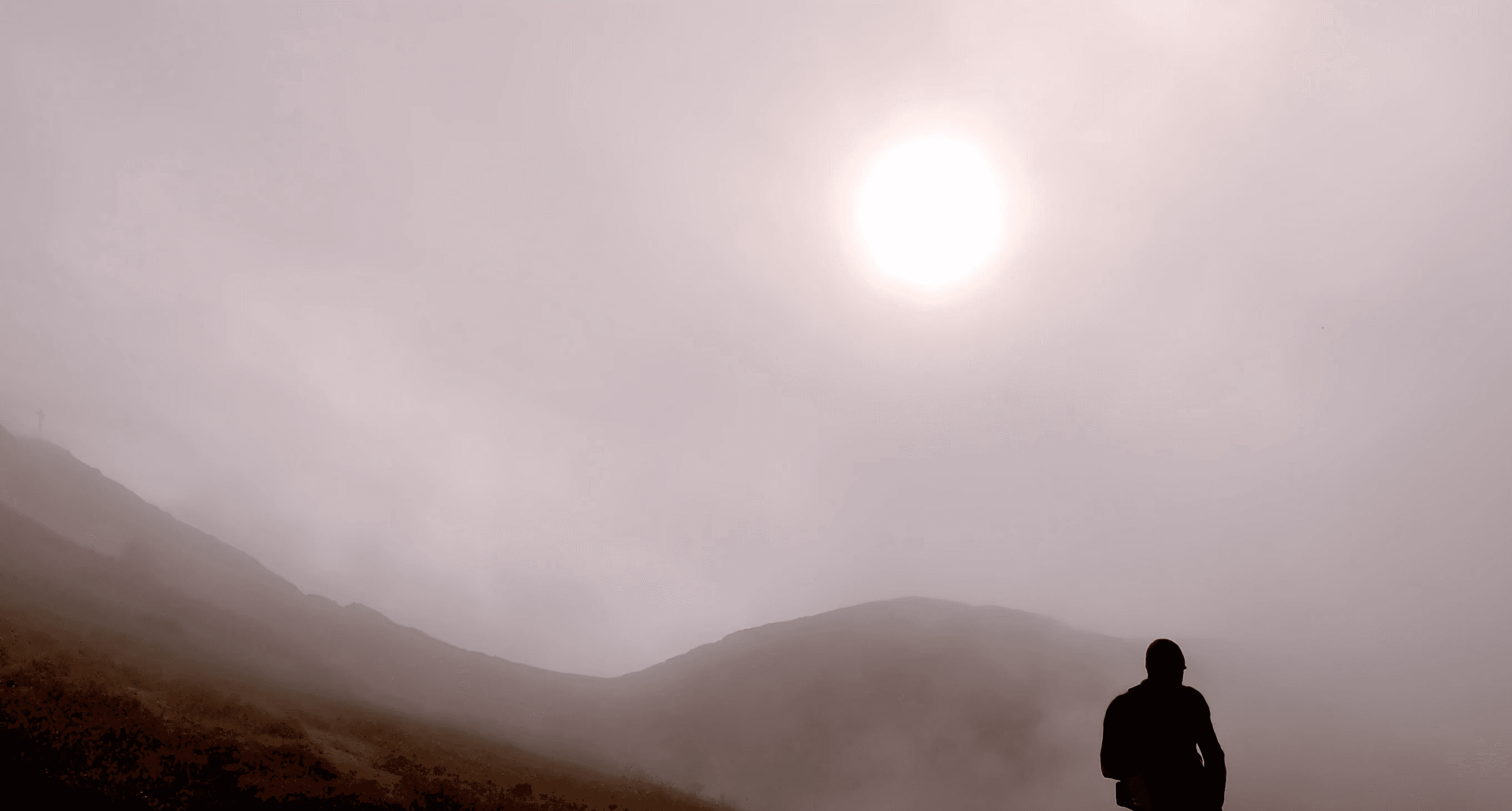 A silhouetted figure stands in foggy terrain, with a bright sun against a hazy sky in the background.