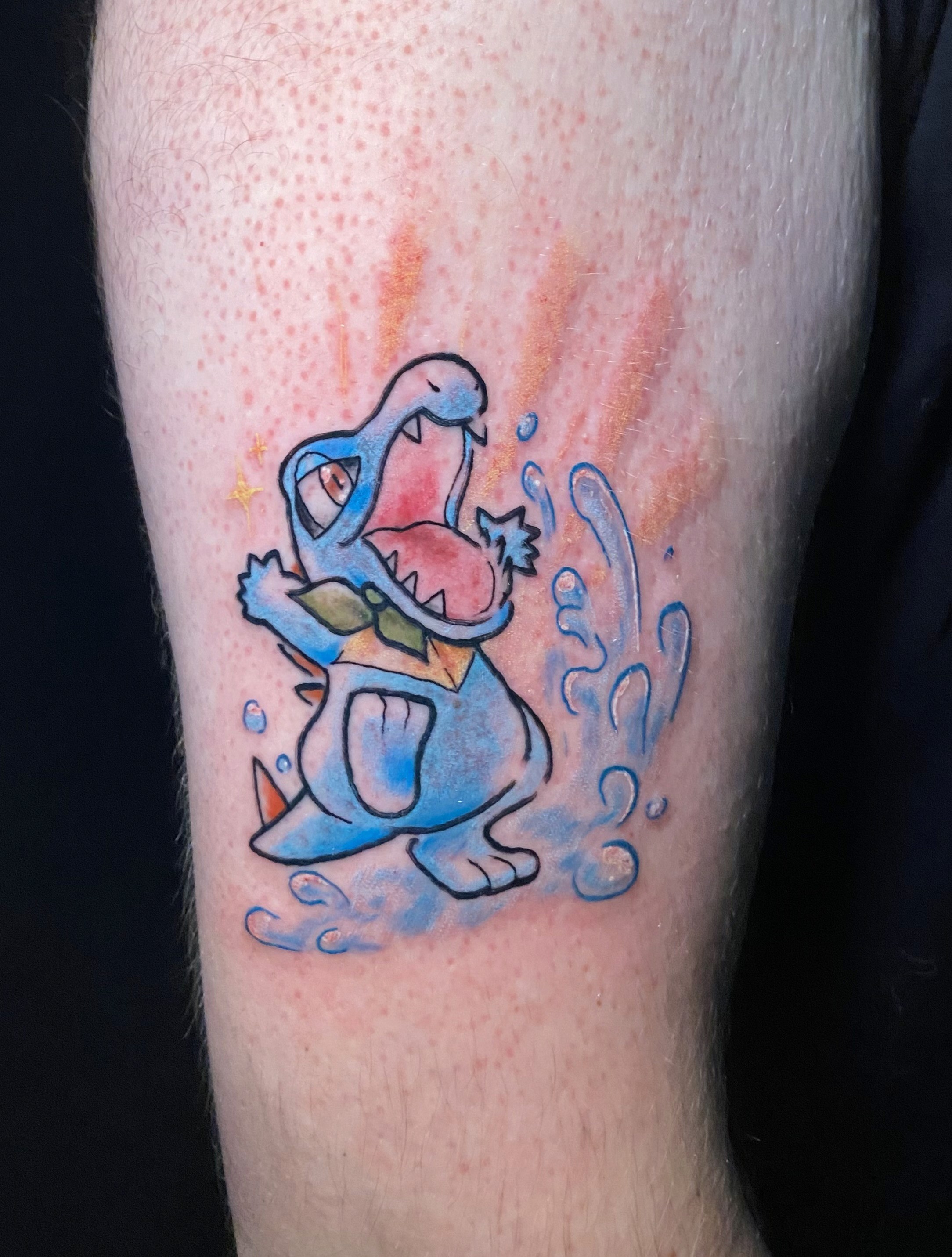 A vibrant tattoo of a chainsaw-like creature on a person's forearm. The tattoo is detailed with shades of orange, black, teal, and red.