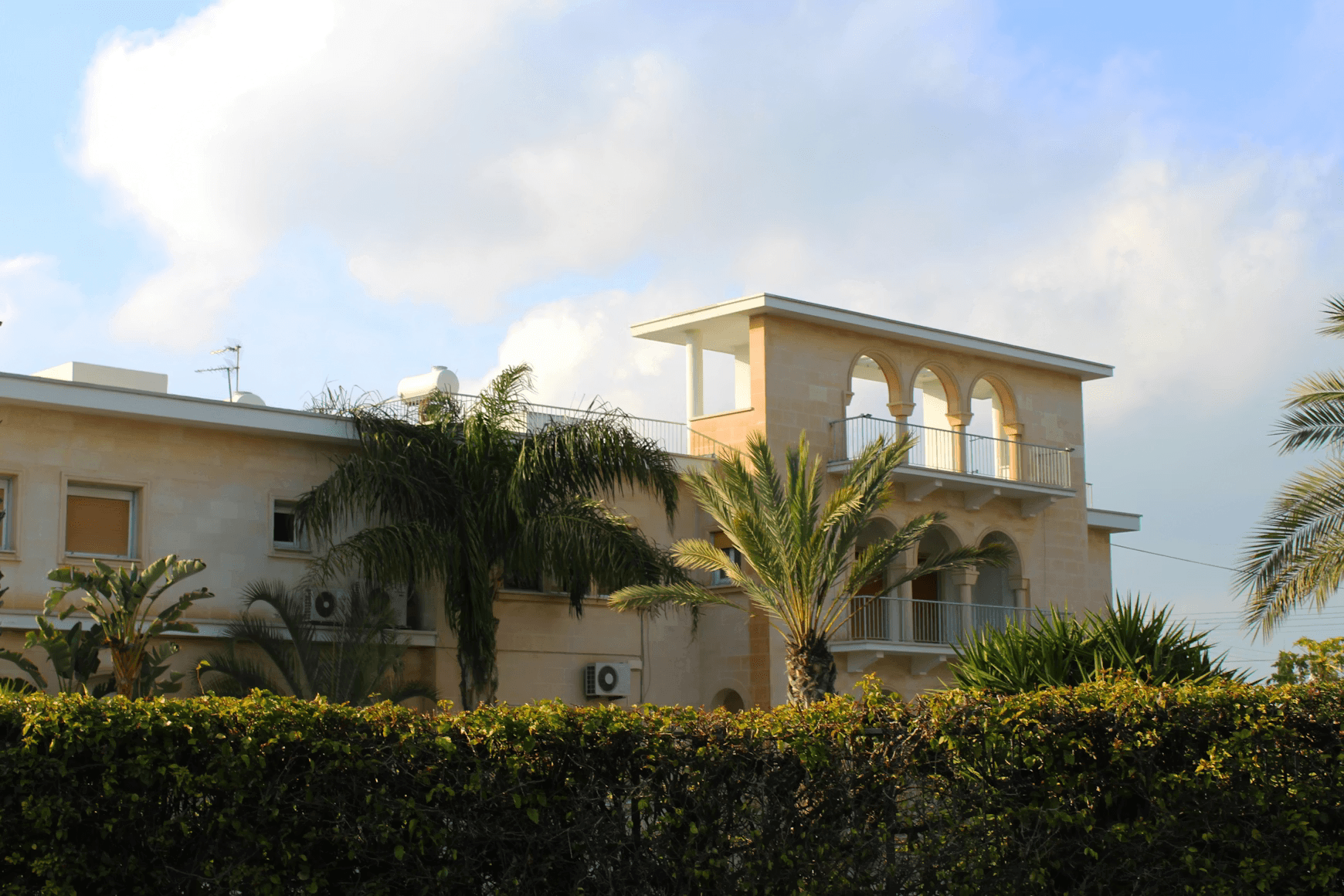 Luxury Mediterranean-style mansion with beige stucco walls, arched tower, palm trees, and manicured hedges under partly cloudy sky.
