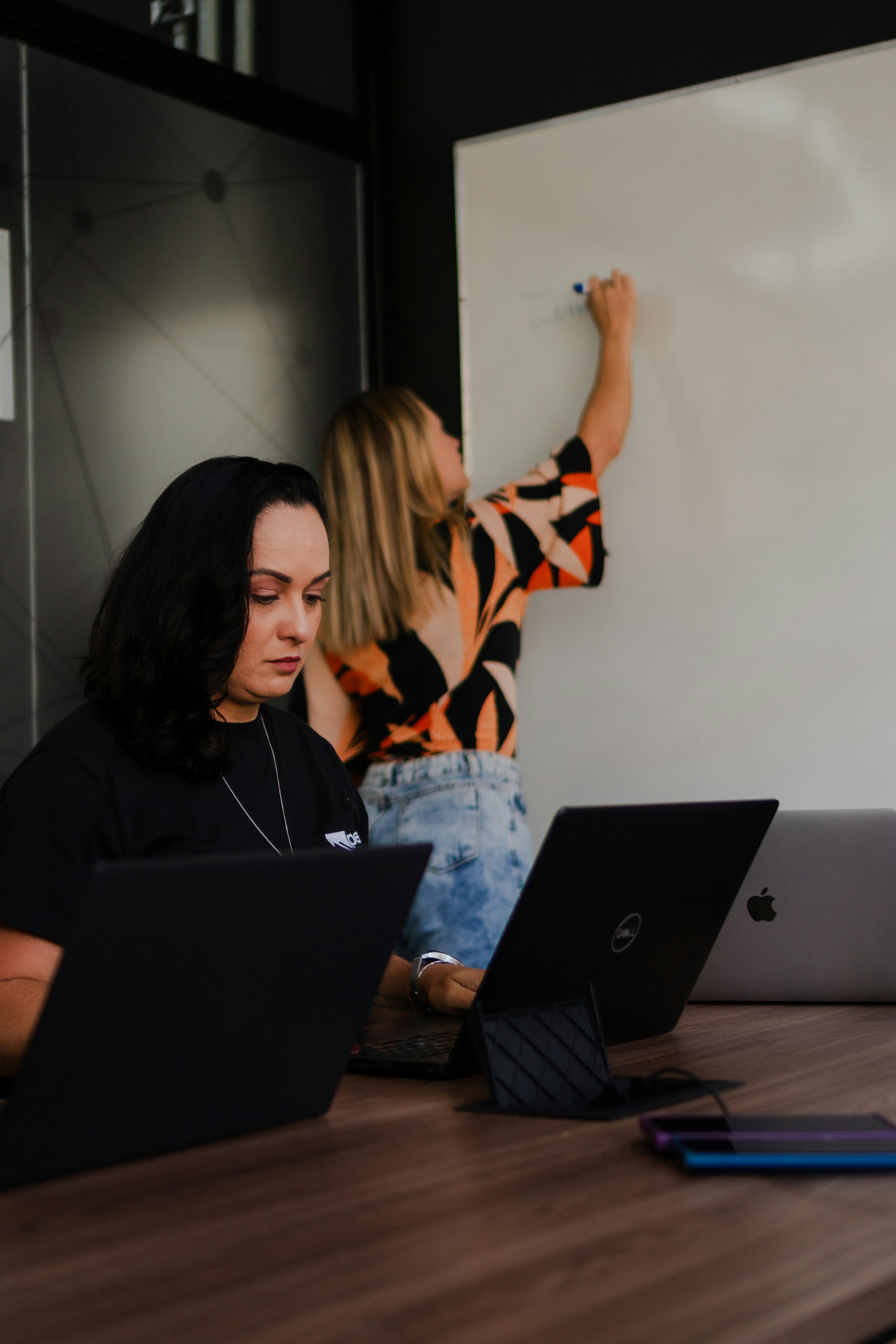 Two women working on laptops near whiteboard.