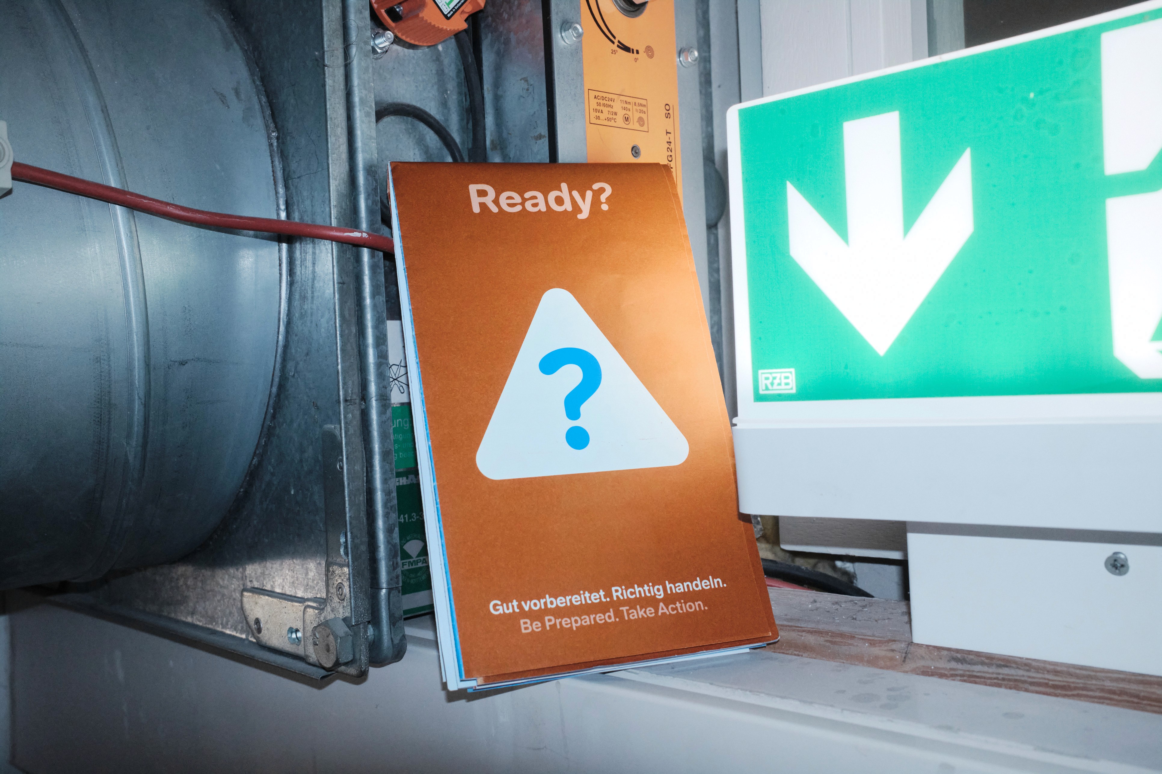 A bronze-colored emergency preparedness booklet titled "Ready?" stands propped between industrial metal equipment and a green-and-white emergency exit sign. The booklet features a large white triangle with a blue question mark and the bilingual slogan: "Gut vorbereitet. Richtig handeln. Be Prepared. Take Action."