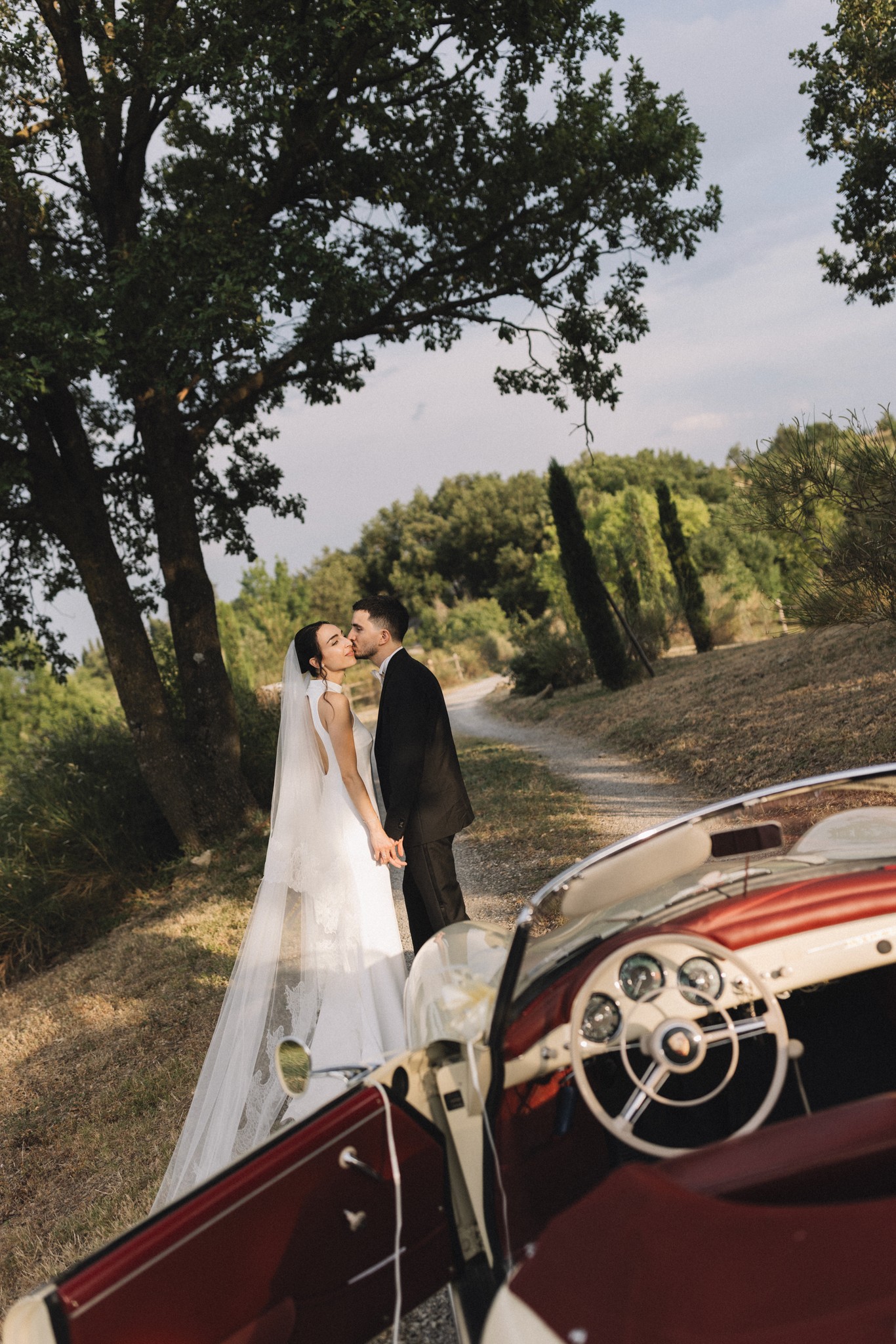 Sposi si baciano accanto a cabriolet d'epoca rossa tra alberi al Parco di Montebello, velo da sposa al vento
