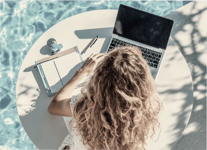 Poolside workspace with a woman using a laptop and notebook on a round white table.