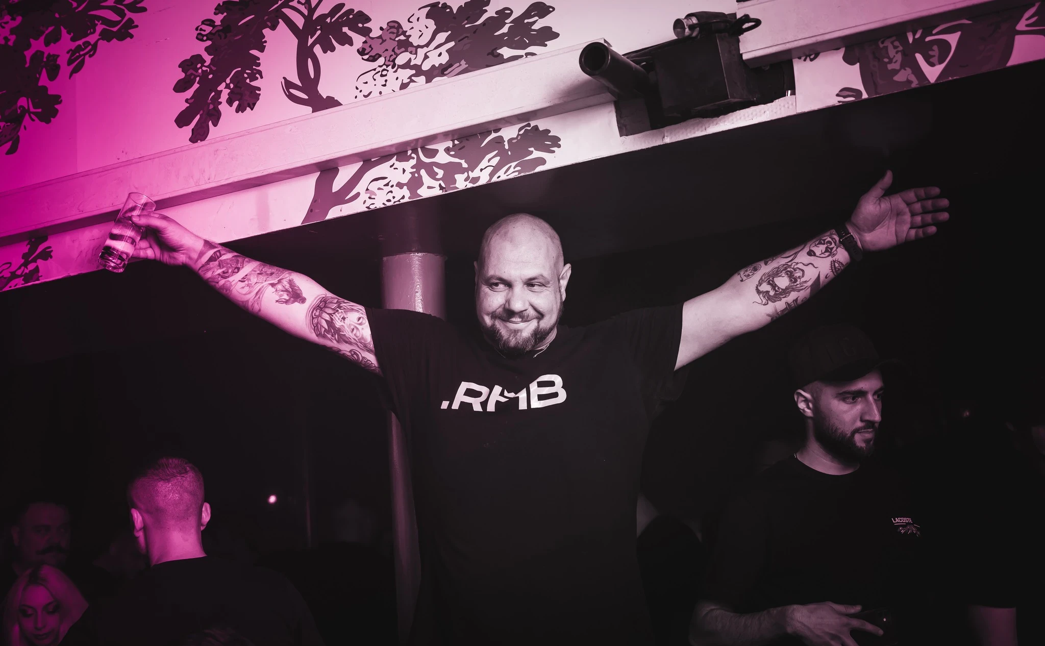 Provocateur, a man with a beard and tattoos on both arms stands with his arms spread wide, smiling and holding a drink in a club. He is wearing a black t-shirt with ".RHB" on it.