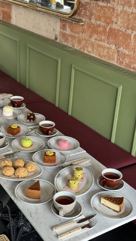 A cozy cafe scene features a row of small plates with an assortment of colorful pastries, cakes, and cups of tea on a white marble table against a green and brick backdrop.