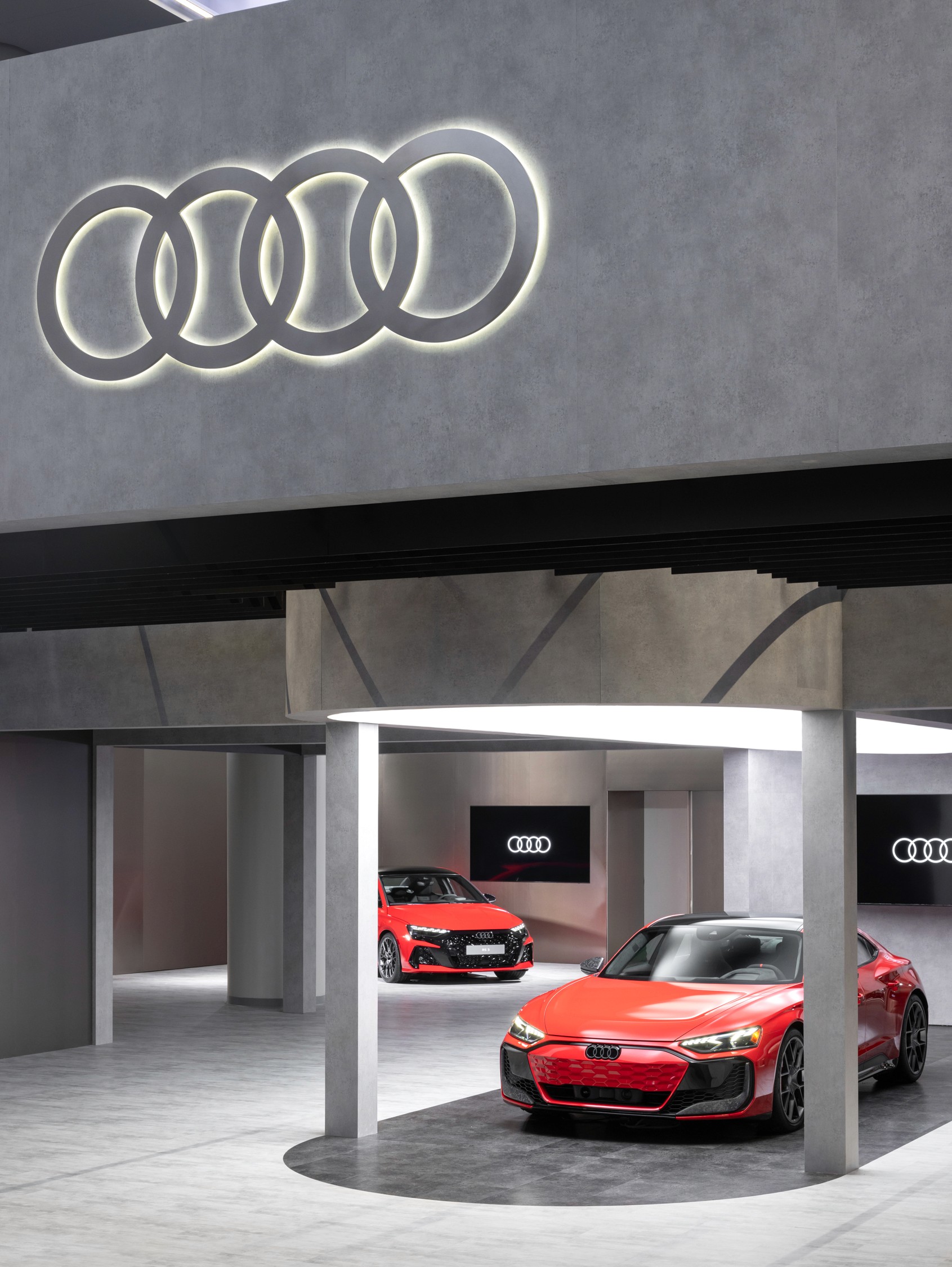 Two sleek, red Audi cars are showcased in a modern showroom under a large illuminated Audi logo, conveying luxury and innovation.