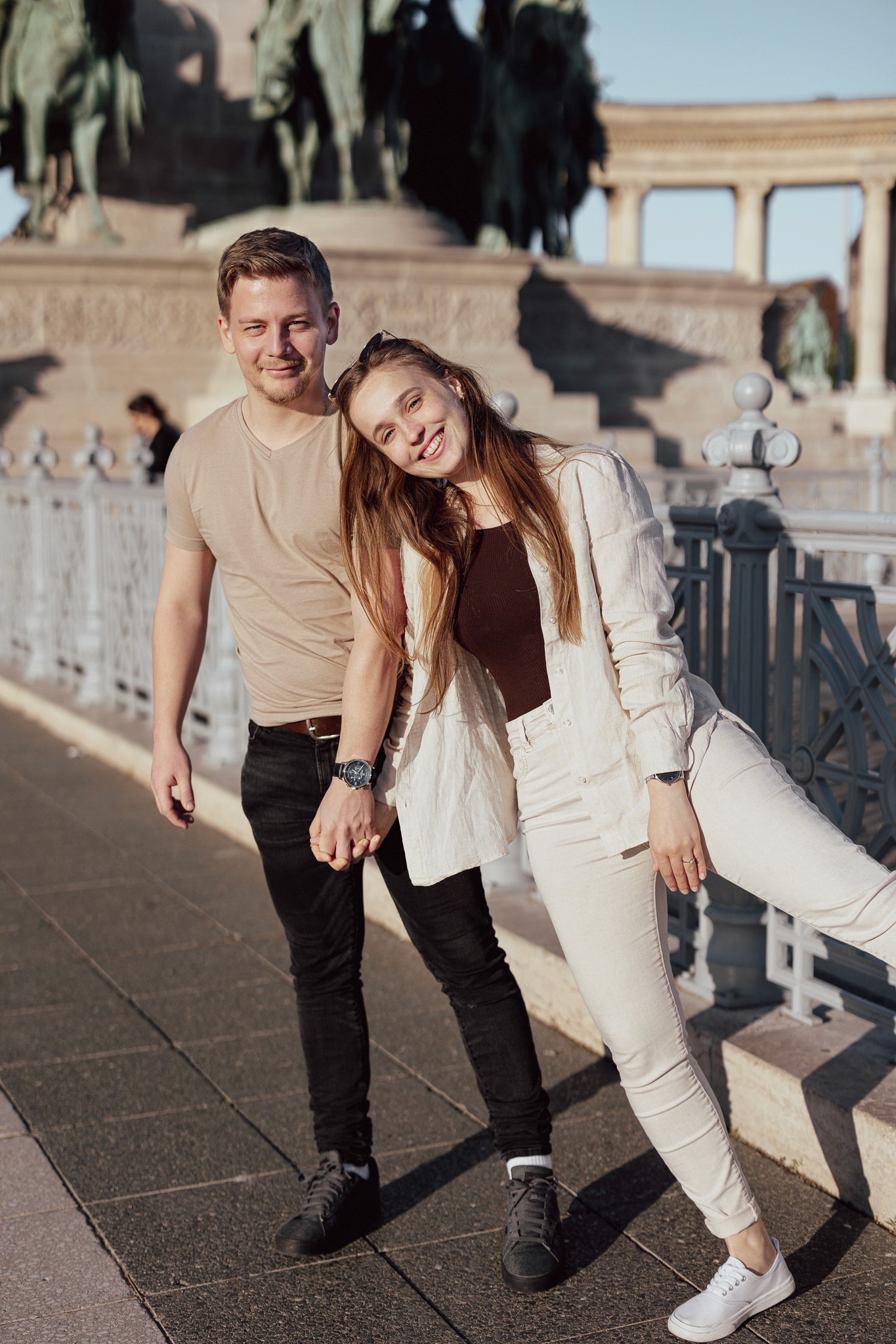 Egy fiatal pár kézen fogva áll a Hősök terén, Budapest ikonikus szoborcsoportja előtt, mosolyogva, napfényben.