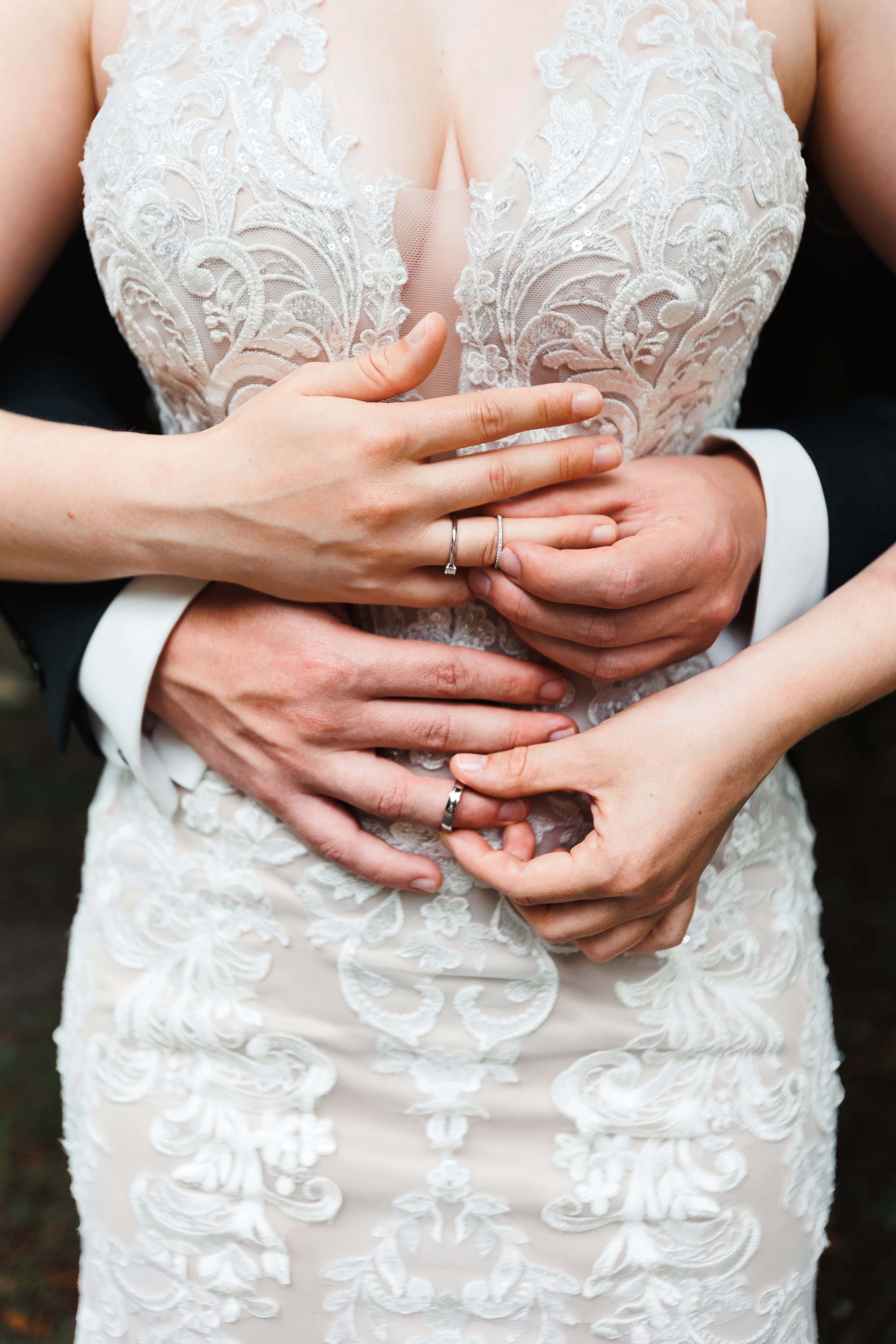 Elegáns esküvői fotó Nagyatádon. A pár összefonódó kezekkel mutatja jegygyűrűiket, a menyasszony elegáns, virágmintás csipke részletekkel díszített fehér ruhában