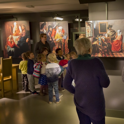 Children and adults observe paintings in a museum, with detailed artworks depicting historical scenes on the walls.