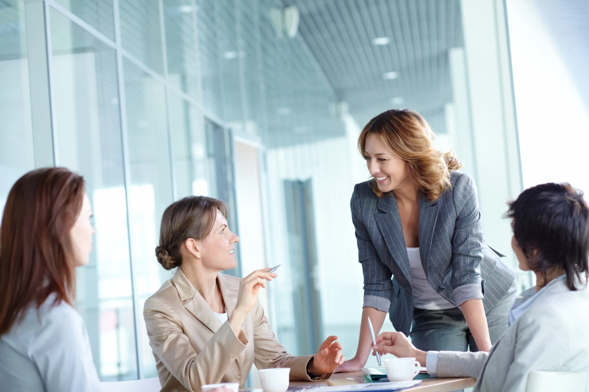 A imagem mostra um grupo de mulheres em um ambiente corporativo, aparentemente em uma reunião. Elas estão bem vestidas em trajes formais e parecem engajadas em uma conversa descontraída, mas produtiva. Uma das mulheres, em destaque, está de pé, inclinada sobre a mesa, sorrindo para as colegas, transmitindo uma sensação de liderança e colaboração. O ambiente é bem iluminado, com paredes de vidro, o que reforça uma atmosfera moderna e profissional. Essa imagem é representativa de mulheres em papéis de liderança e em um ambiente de trabalho colaborativo.