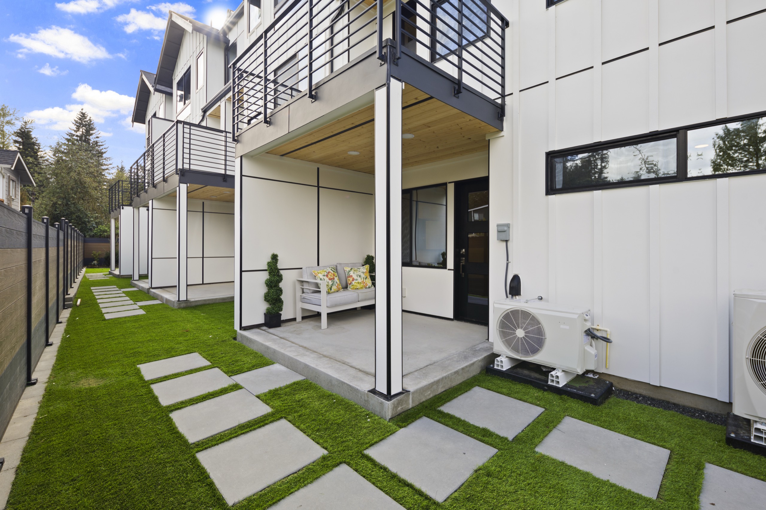 Landscaped backyard of a luxury townhome with artificial grass, concrete stepping stones, covered patio with bench, upper balcony, and modern white exterior