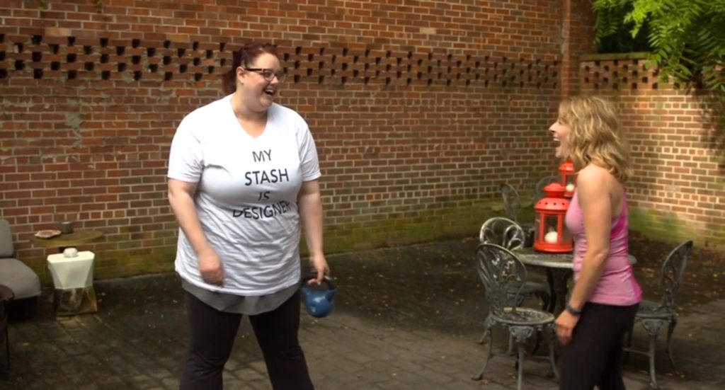 Two women share a joyful moment in a brick-walled courtyard, with one holding a kettlebell and the other smiling, amidst outdoor furniture and a red lantern.