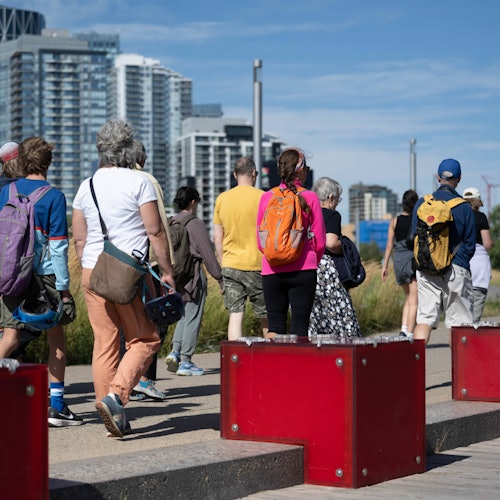 Group of people walking outdoors with backpacks, surrounded by city buildings and red blocks.