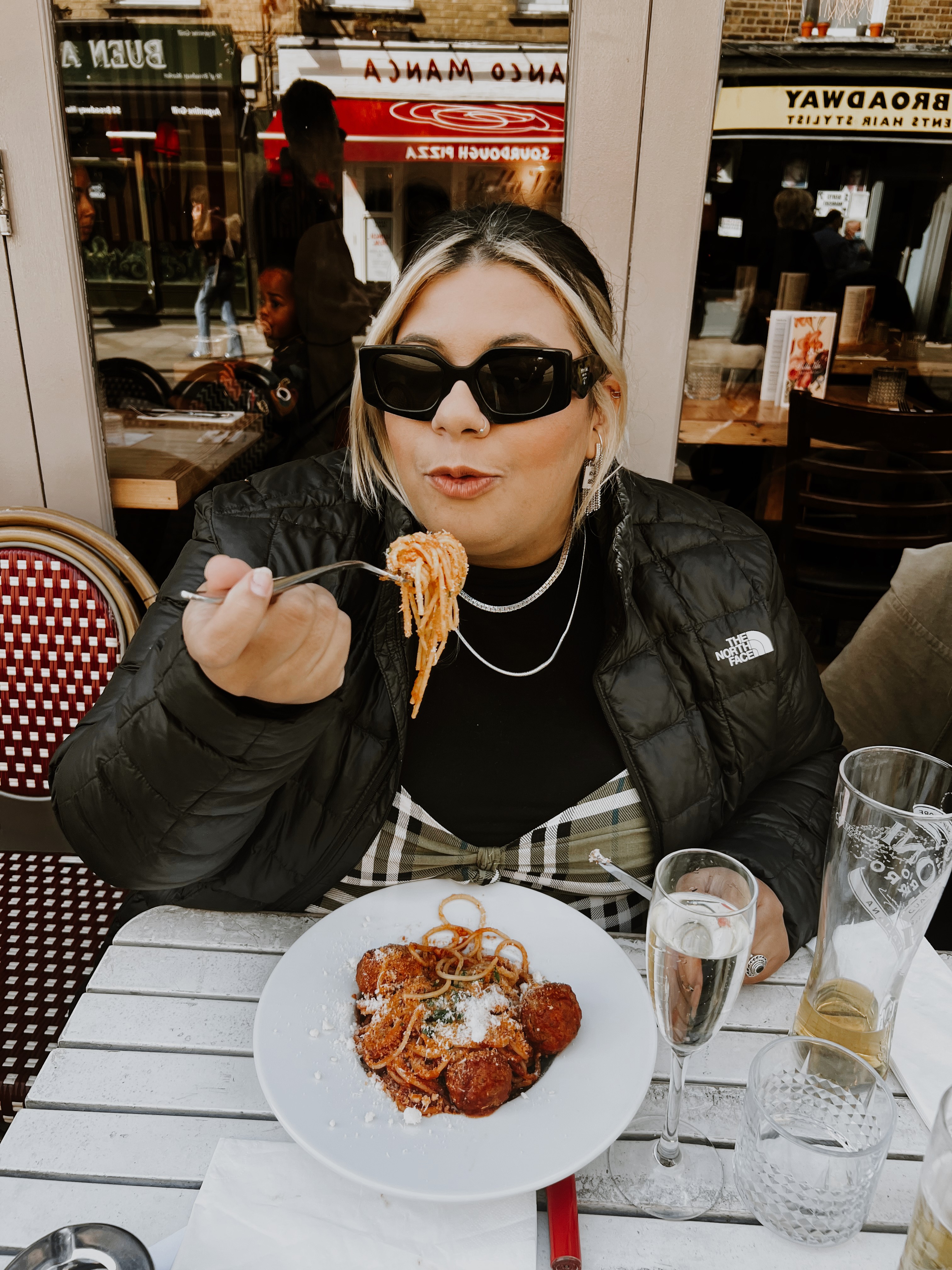 A young person with curly hair smiles at the camera while sitting in front of a laptop against a dark background.