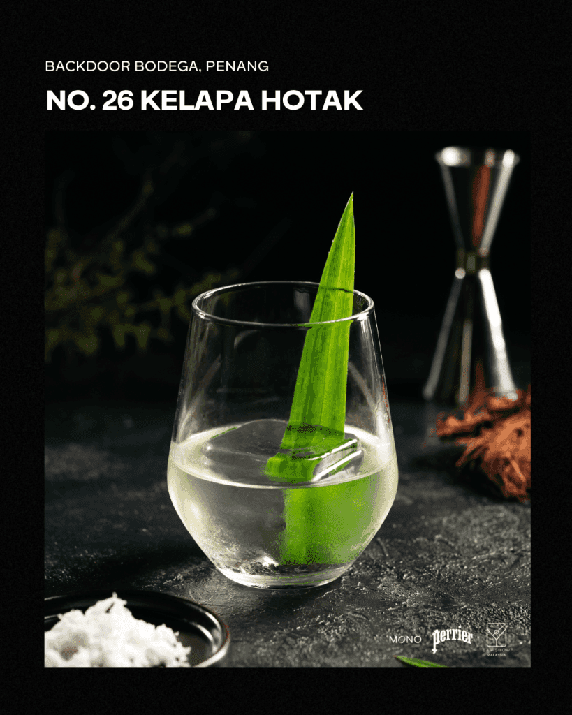 A clear cocktail named 'Kelapa Hotak' served in a glass, garnished with a long pandan leaf. The background features dark stone and some coconut shavings.