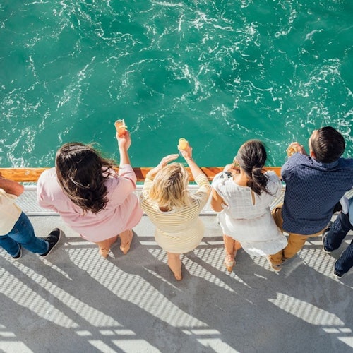 Five people standing on a boat deck holding drinks, looking at the turquoise water below.