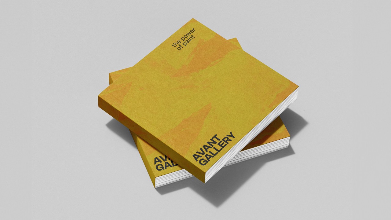 Two square yellow-ochre exhibition catalogues stacked on a pale grey surface, one titled “the power of paint” and both stamped “AVANT GALLERY” at the corner.