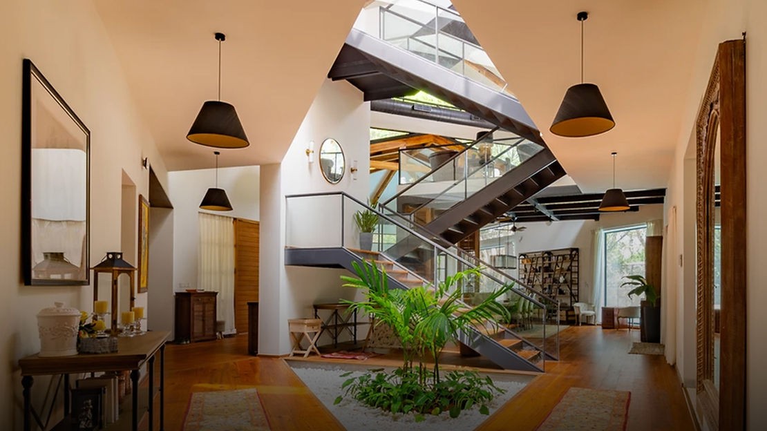 A foyer with brown wood floors and two rugs on either side. The ceiling above is open and lets daylight in. A triangular garden in the middle of the foyer with green plants. Behind the plants is a flight of stairs with glass railings. Lamps hang from the ceiling, and there are framed pictures on the white walls. Shelves along the walls have ornaments and decorations.
