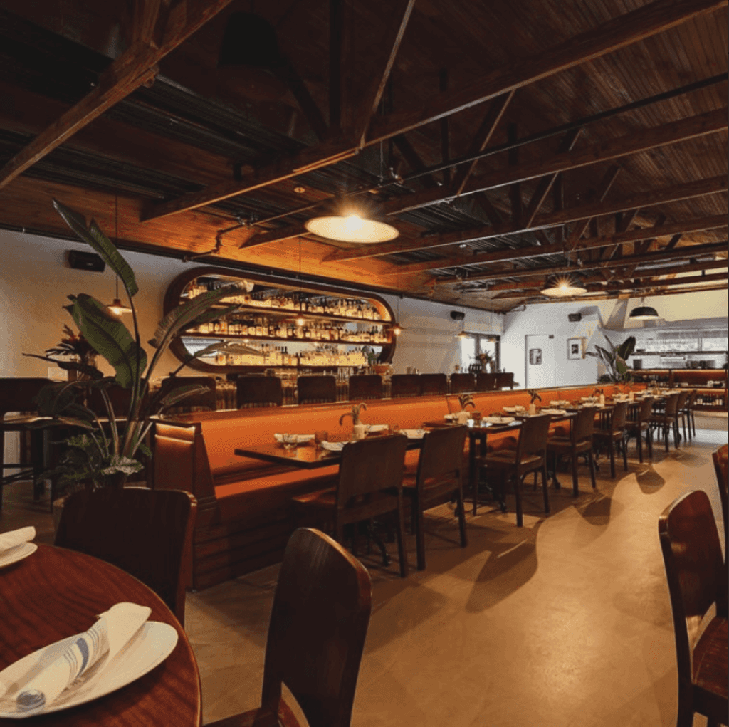 Interior of Macchialina restaurant featuring wooden ceilings, warm lighting, and a bar with bottles and plants creating an inviting atmosphere.