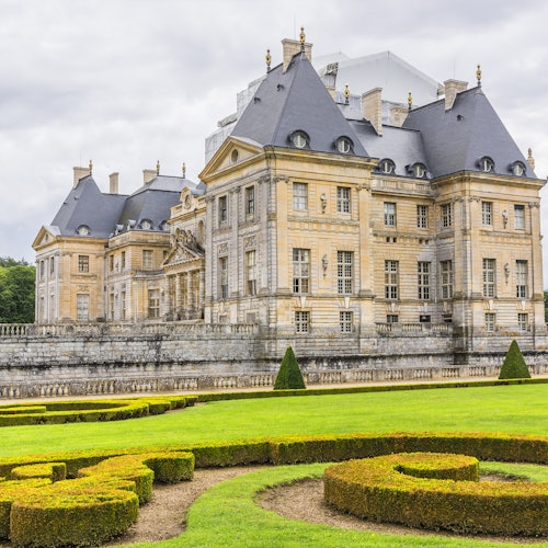 Изящный классический каменный замок с шиферными крышами и ухоженными садами, в переднем плане которых расположены изысканные живые изгороди.