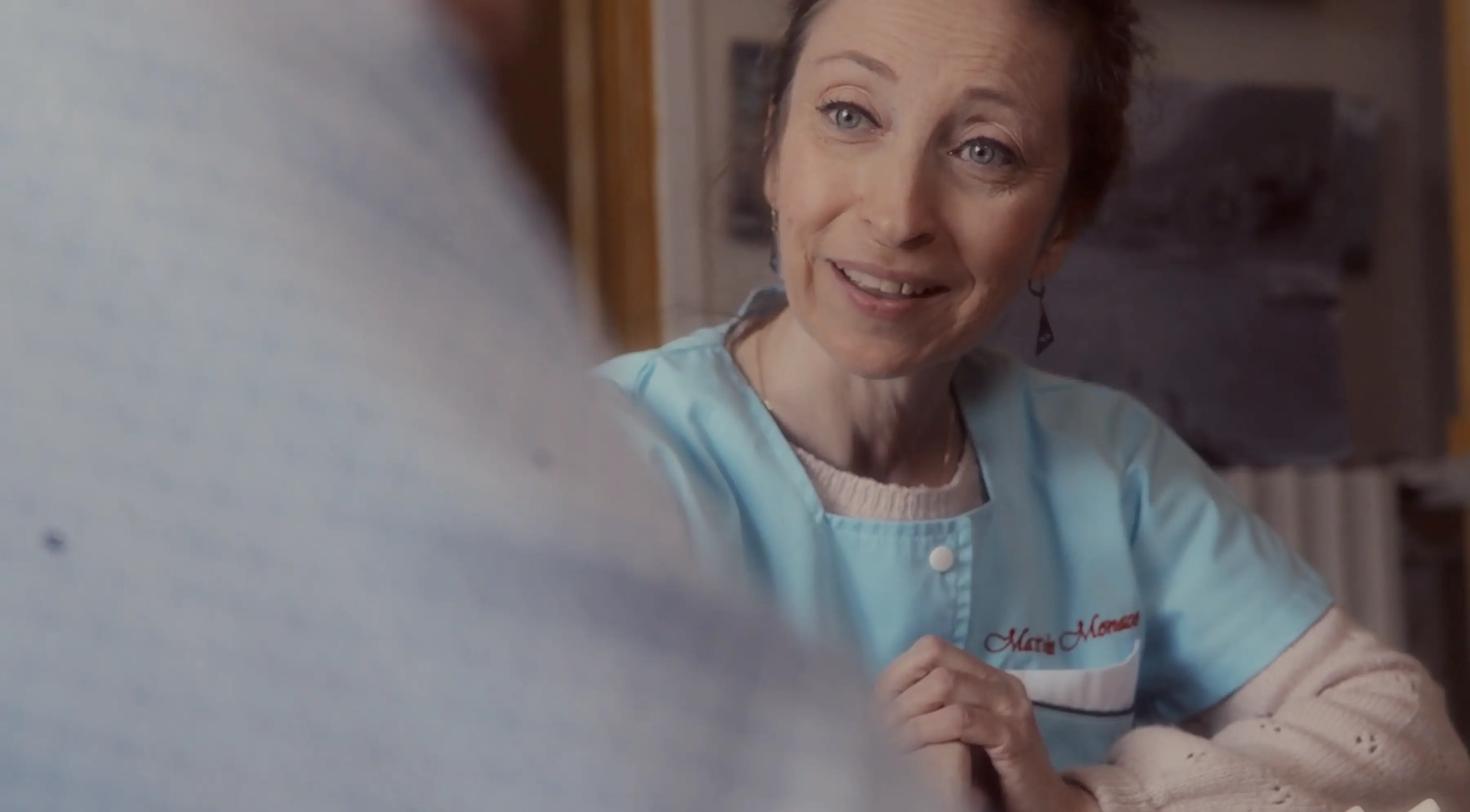 A smiling healthcare worker wearing a blue uniform with a name tag sits attentively with clasped hands, engaging in conversation, in a warmly lit room.