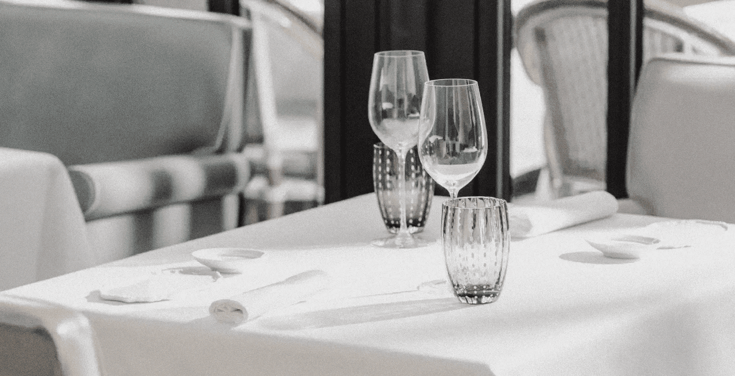 A sunlit restaurant table set with crystal glasses and white linens — minimal, sophisticated, and ready for an intimate culinary experience.