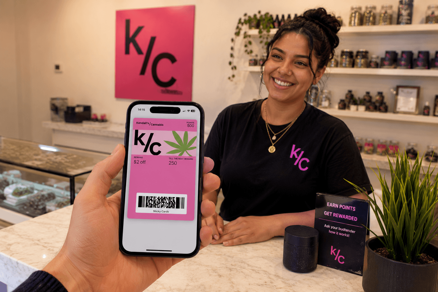 A realistic cannabis dispensary scene showing a customer holding a smartphone displaying a bright pink digital loyalty wallet pass card with a cannabis leaf graphic, reward details, and barcode. Across the counter, a smiling dispensary employee stands in a modern retail setting with shelves of products, small plants, and warm lighting in the background. A large pink “K/C” style wall sign reinforces the store branding, while the overall atmosphere feels welcoming, modern, and customer-friendly.
