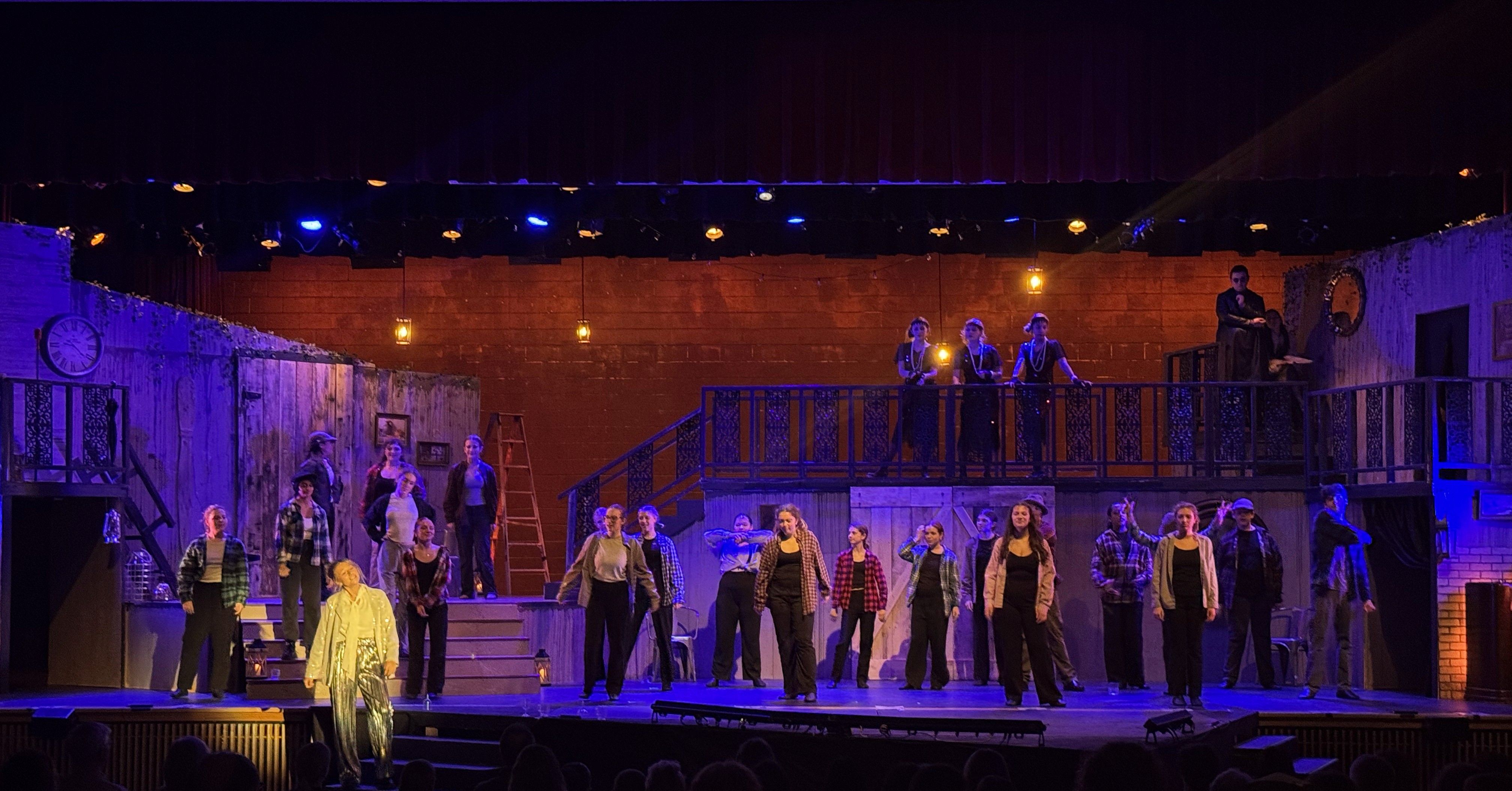 An ensemble performs "Way Down Hadestown" on a grand stage set against a rustic orange backdrop, dancing under blue top lighting while Hermes, in a spotlight, speaks.