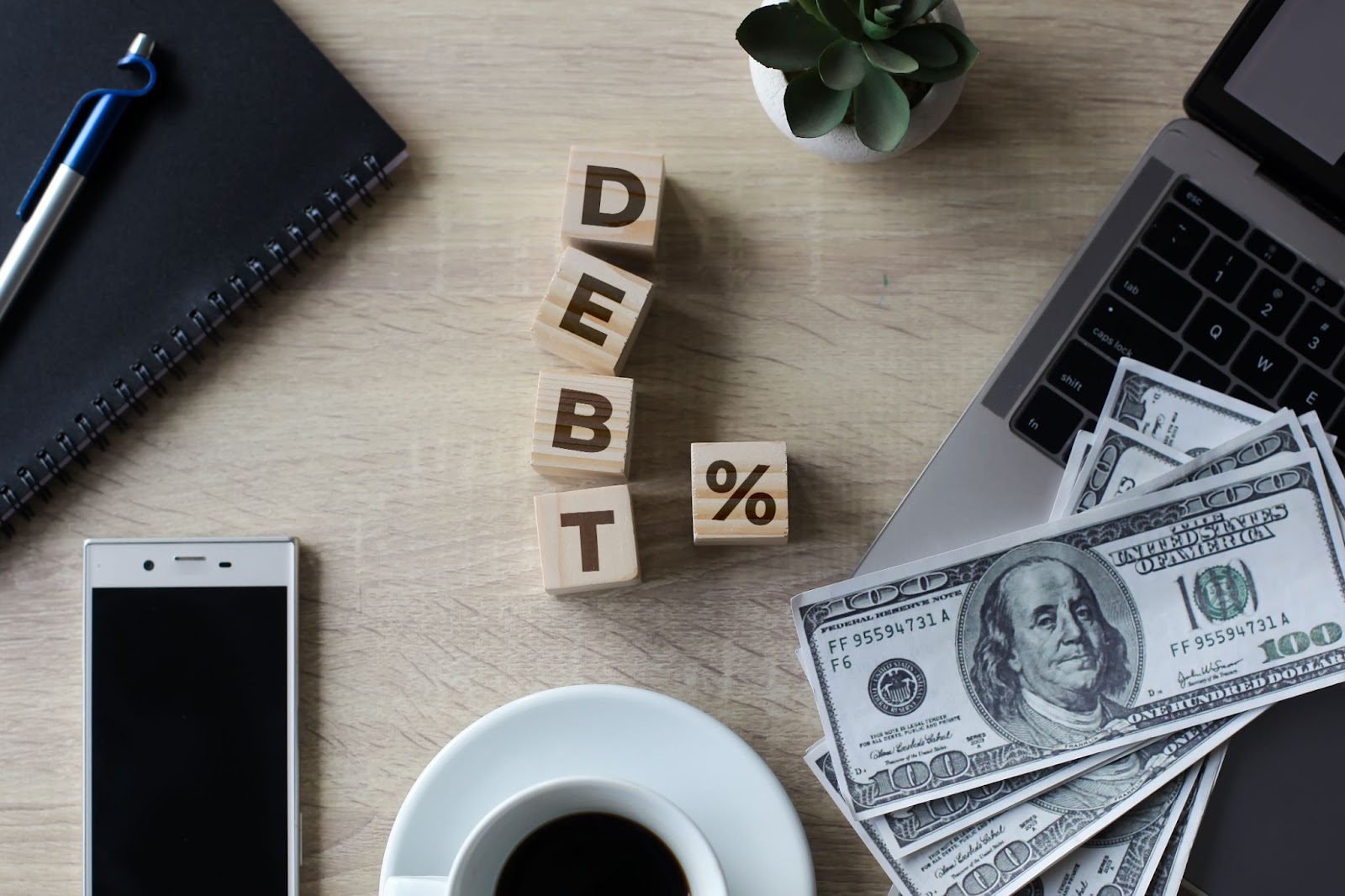 Word “debt” on blocks next to a laptop, cell phone, cup of coffee, and stack of money. 