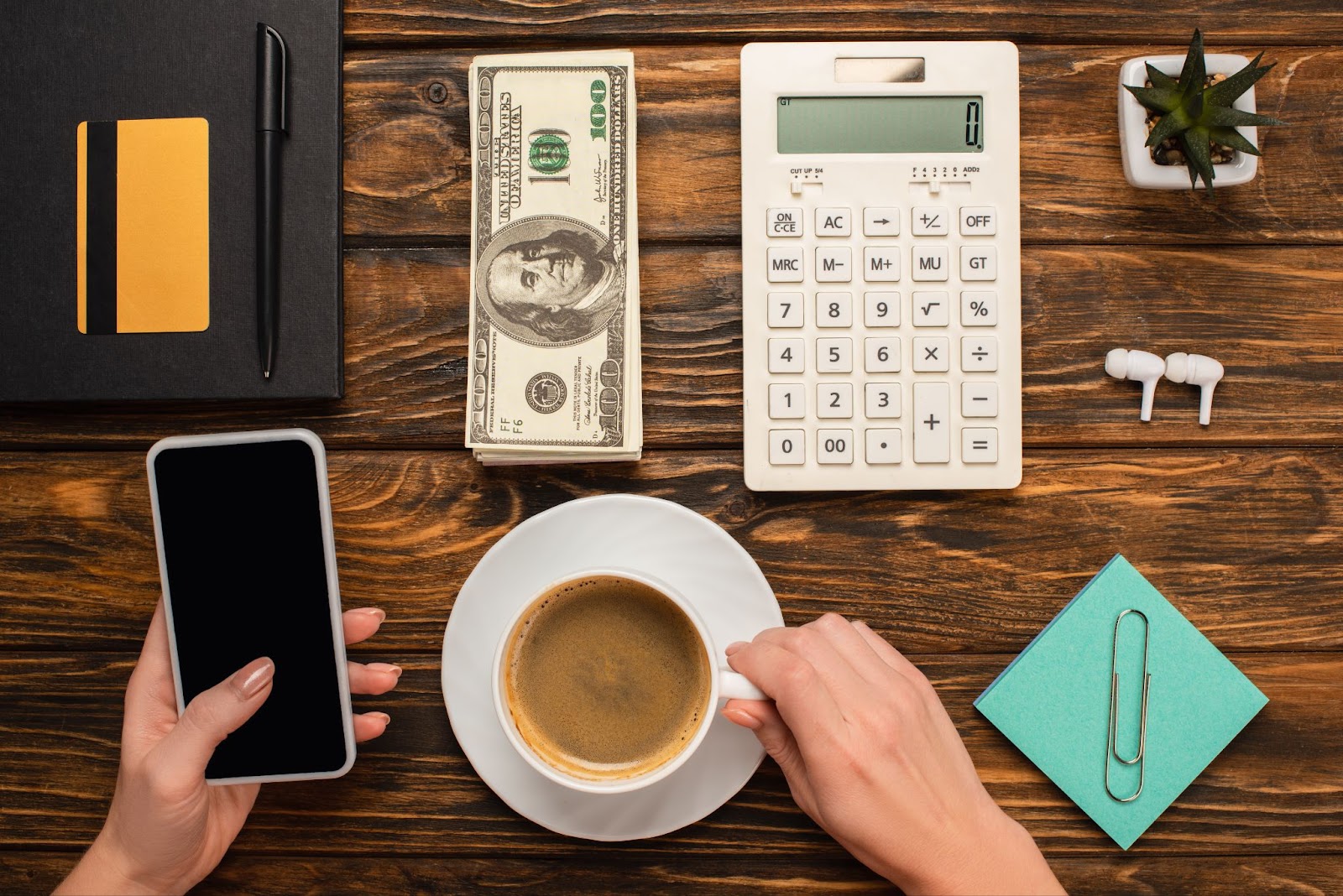 Woman holding a cell phone in her hand next to a cup of coffee, notebook, credit card, pen, stack of money, calculator, plant, notepad, and earbuds. 