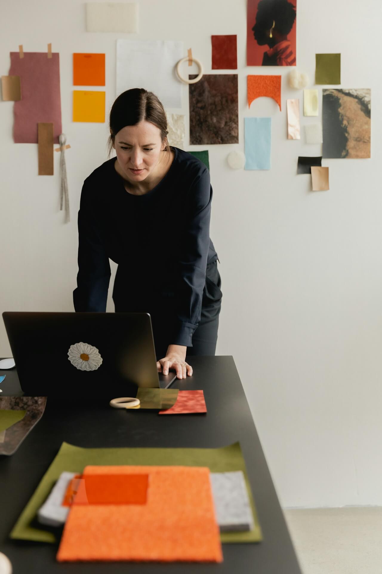 A woman in black stands focused over a laptop on a table, surrounded by colorful paper samples pinned to a white wall, creating an artistic, thoughtful atmosphere.
