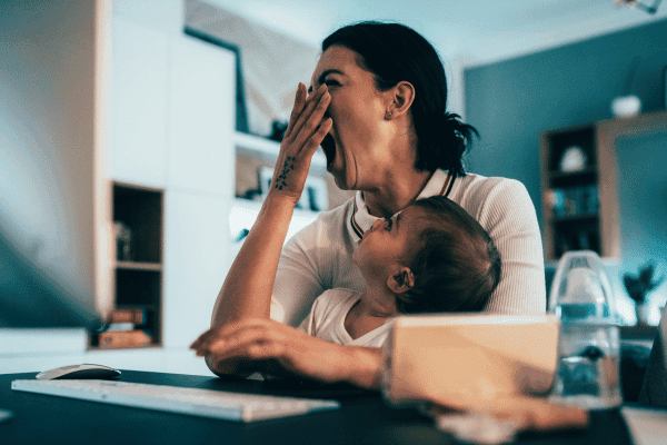Tired mother yawning while holding her baby at home, highlighting the importance of health insurance for busy families.