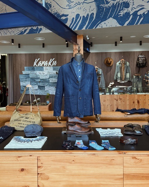 A display in a clothing store featuring a denim blazer on a mannequin, with casual shirts, shoes, and accessories arranged on a table, and a wooden interior design in the background.