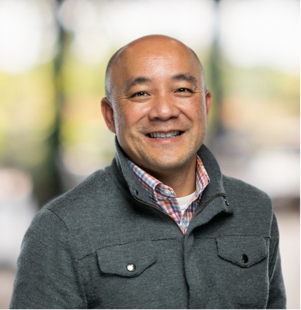A imagem mostra um homem sorridente em uma jaqueta cinza com uma camisa xadrez colorida por baixo, em pé ao ar livre contra um fundo natural suavemente desfocado.