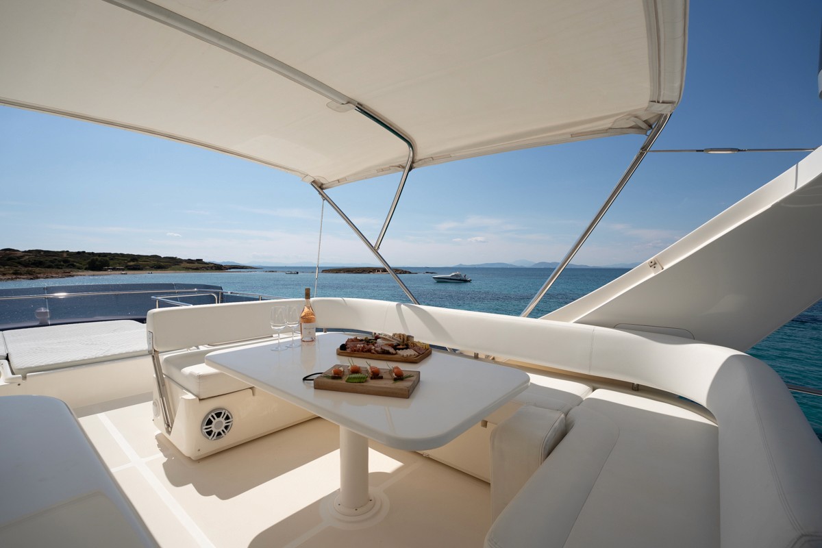Modern white Ferretti 63 yacht with spacious flybridge featuring dining table set with appetizers, anchored in turquoise Aegean waters.