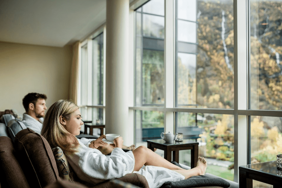 A couple relaxes in a modern, cozy hotel lounge wearing bathrobes, enjoying hot drinks while gazing at a scenic view of a lush, autumnal landscape through large floor-to-ceiling windows.