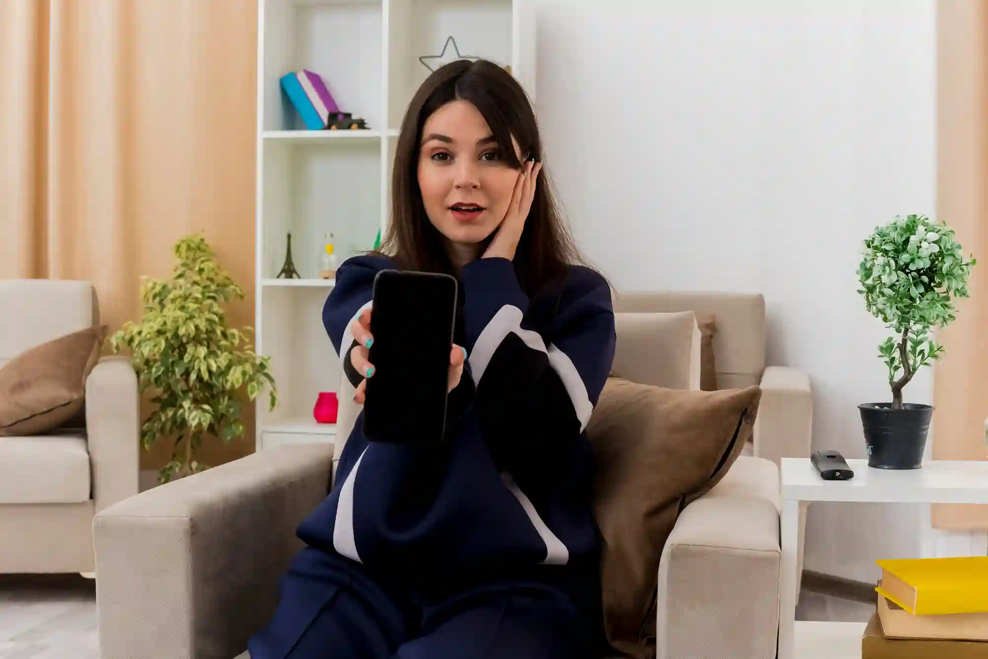 A young woman excitedly showing a blank smartphone screen while sitting in a comfortable, modern home living room.