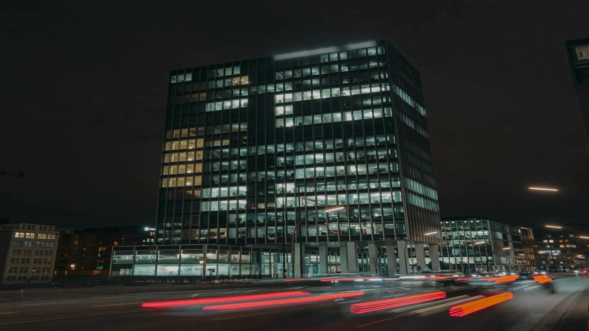 Beleuchtetes Bürogebäude von RITTER Starkstromtechnik bei Nacht. Vor dem modernen Glasgebäude ziehen die Lichtspuren vorbeifahrender Autos über die Straße.